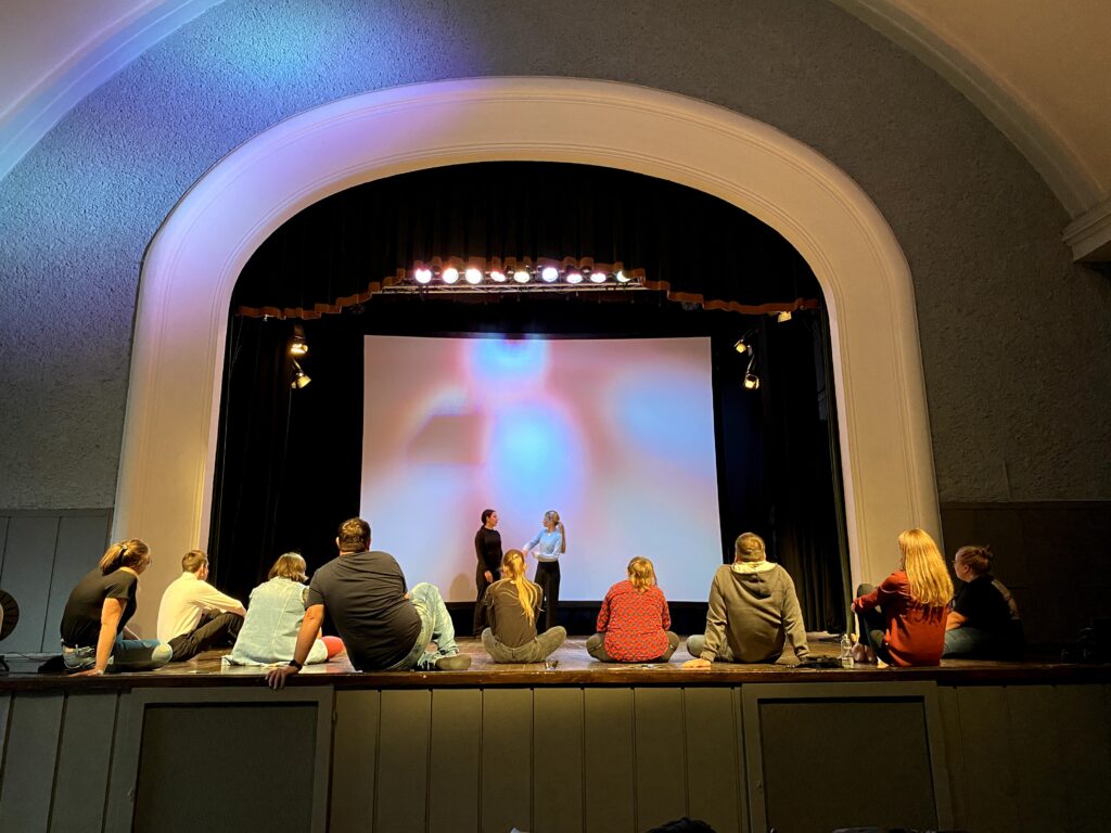 Uczestnicy warsztatów aktorskich siedzą na scenie, w głębi dwie dziewczyny odgrywają scenę